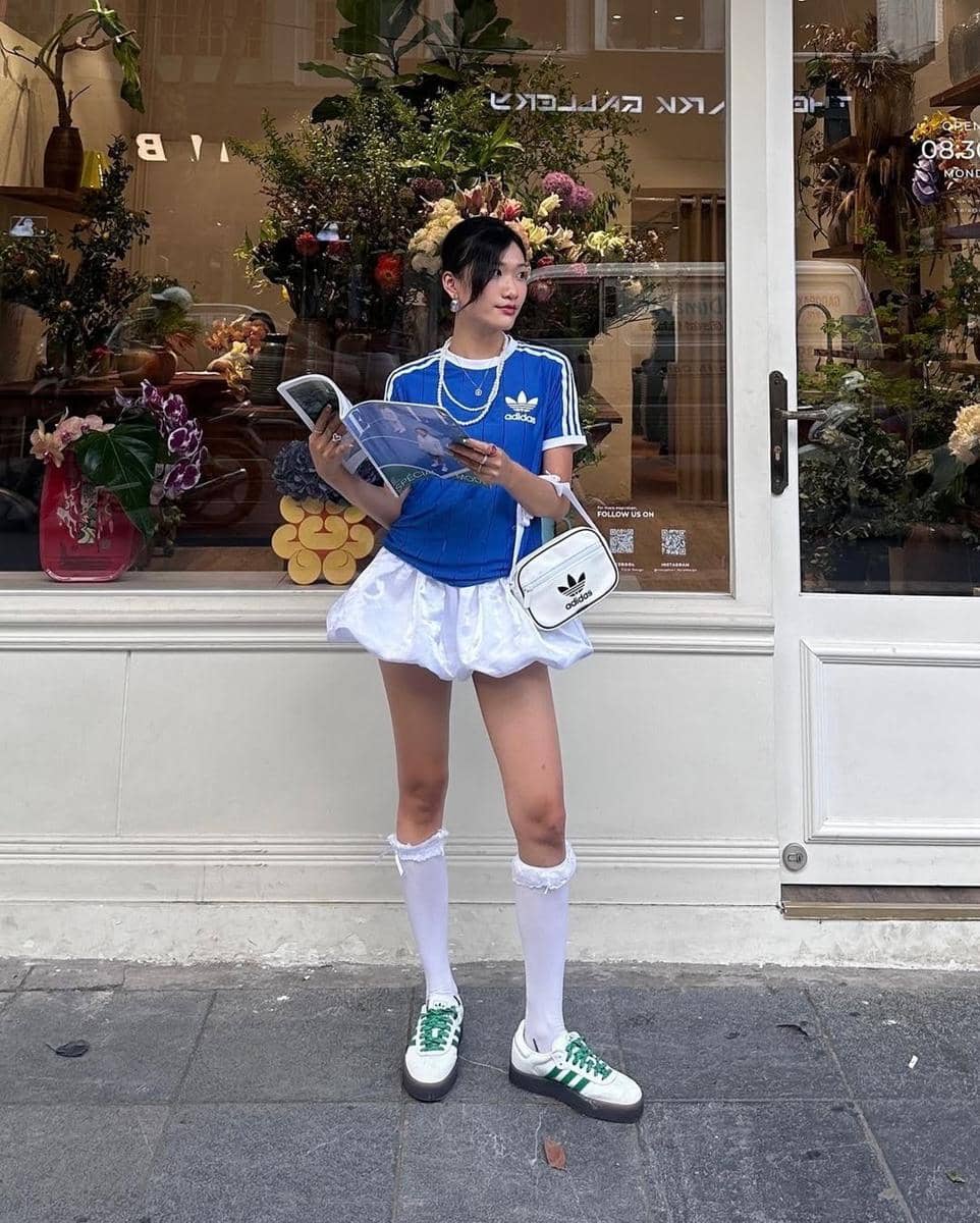 Seorang perempuan berdiri di depan toko bunga sambil memegang majalah, mengenakan kaus biru Adidas, rok putih, dan tas selempang kecil.