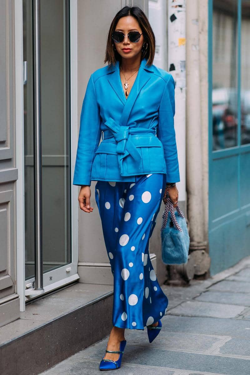 Seorang wanita berjalan di trotoar mengenakan blazer biru dengan ikat pinggang, celana satin biru bermotif polkadot putih, dan tas berbulu biru.