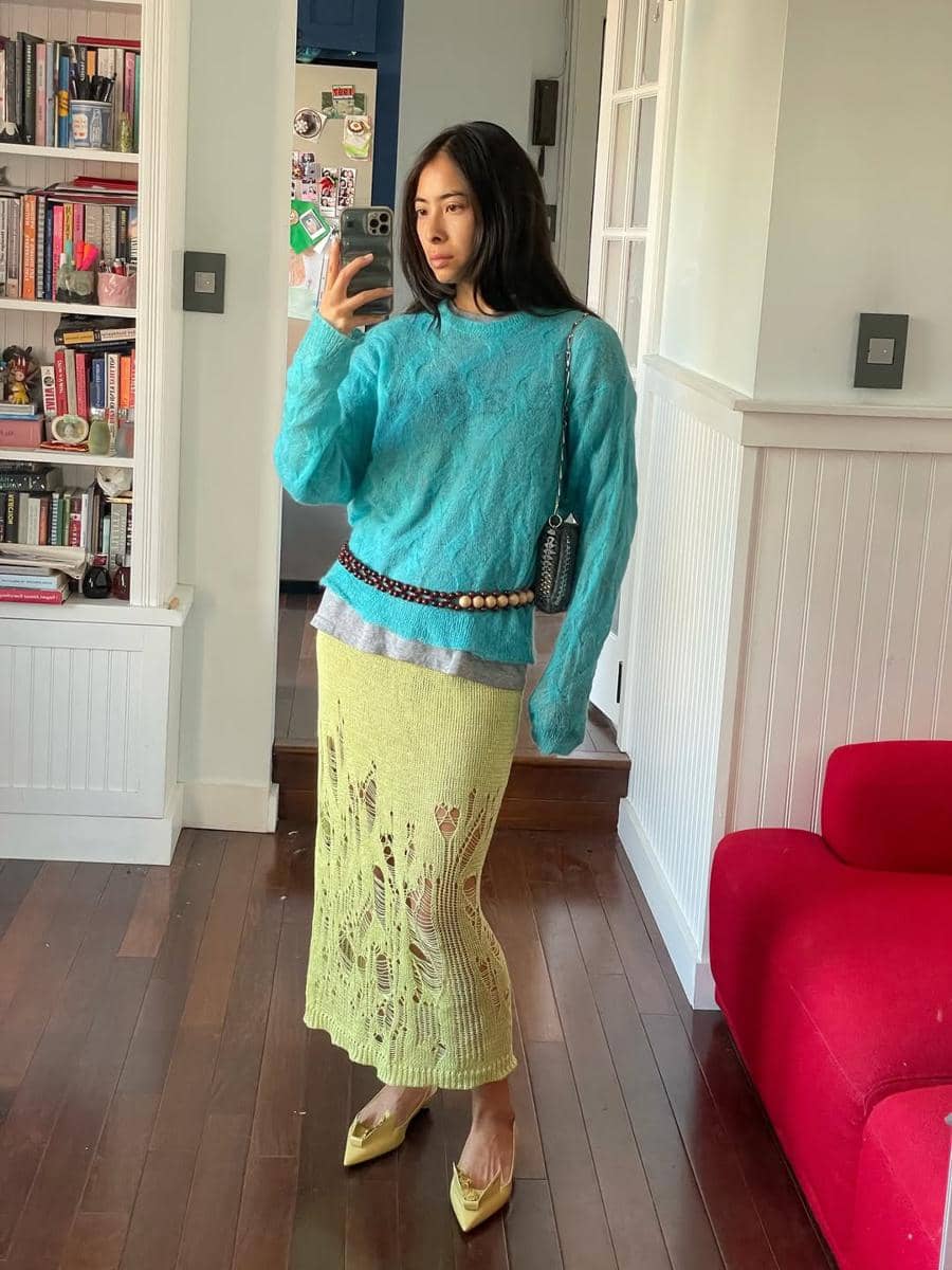 Seorang wanita berpose di depan cermin mengenakan sweater biru muda, rok rajut hijau muda, dan sepatu hak kuning di ruang tamu.