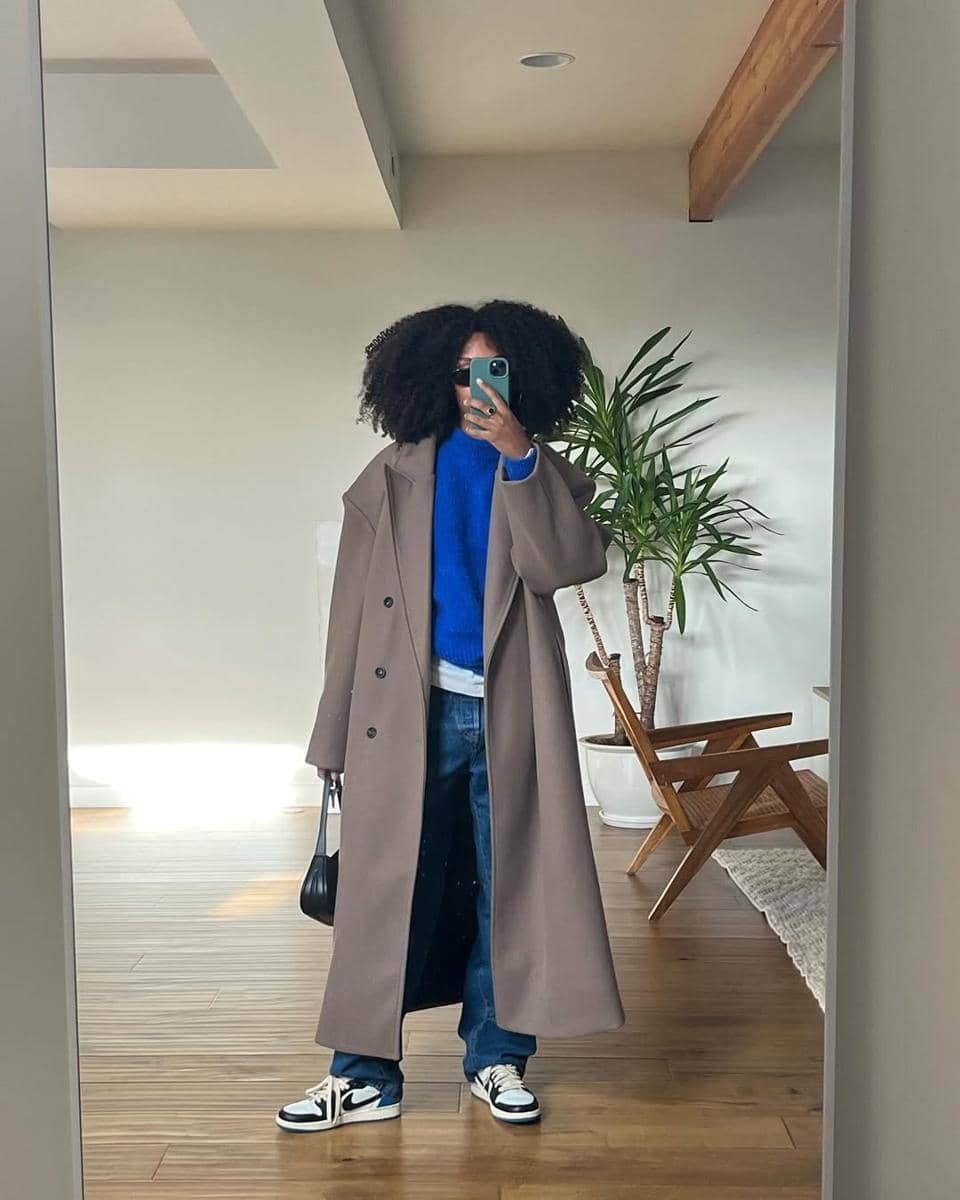 Seorang wanita berpose di depan cermin mengenakan mantel panjang abu-abu, sweater biru, celana jeans longgar, dan sepatu sneakers.