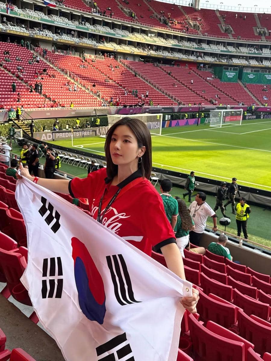Kwon Eunbi mengenakan kaus merah memegang bendera Korea Selatan di stadion sepak bola dengan tribun berwarna merah.