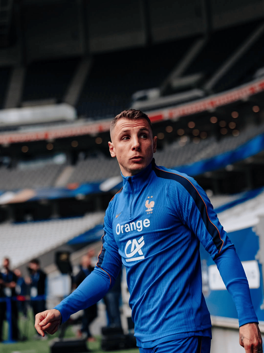 Lucas Digne mengenakan jersey latihan biru Timnas Prancis di stadion dengan latar tribun kosong.