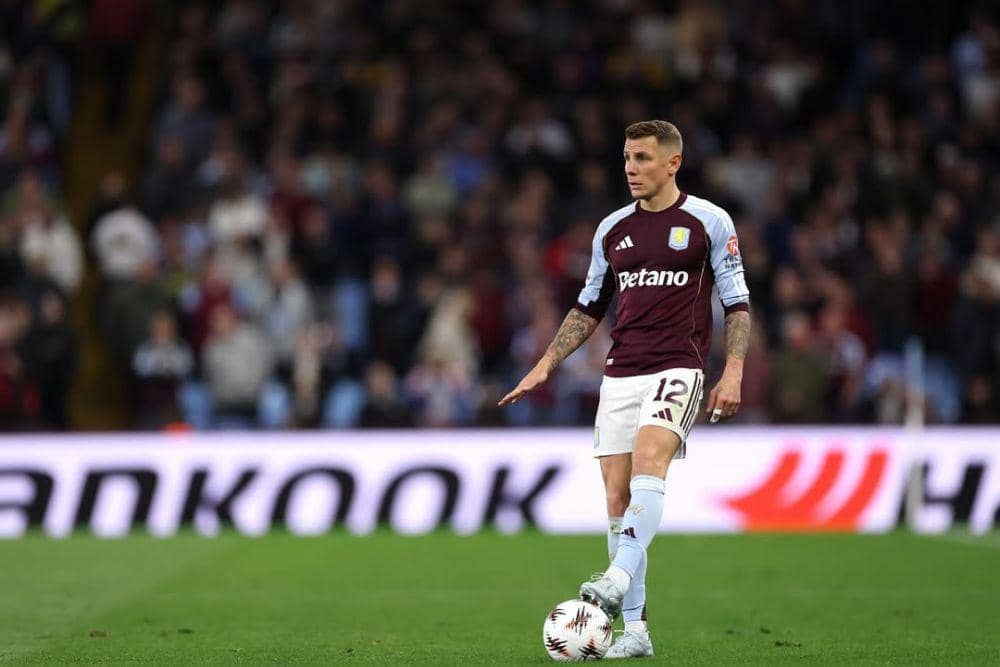 Pemain Aston Villa, Lucas Digne, mengenakan seragam bernomor 12 sedang menguasai bola di lapangan dengan latar penonton di stadion.