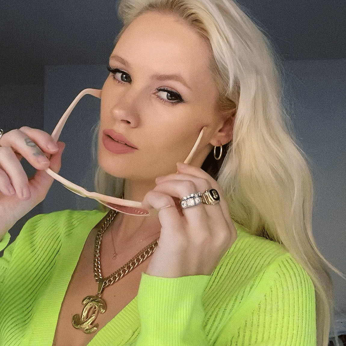 Lauren Bennett yang berambut pirang mengenakan kardigan hijau neon, perhiasan emas, dan memegang kacamata berbingkai krem di depan wajahnya.