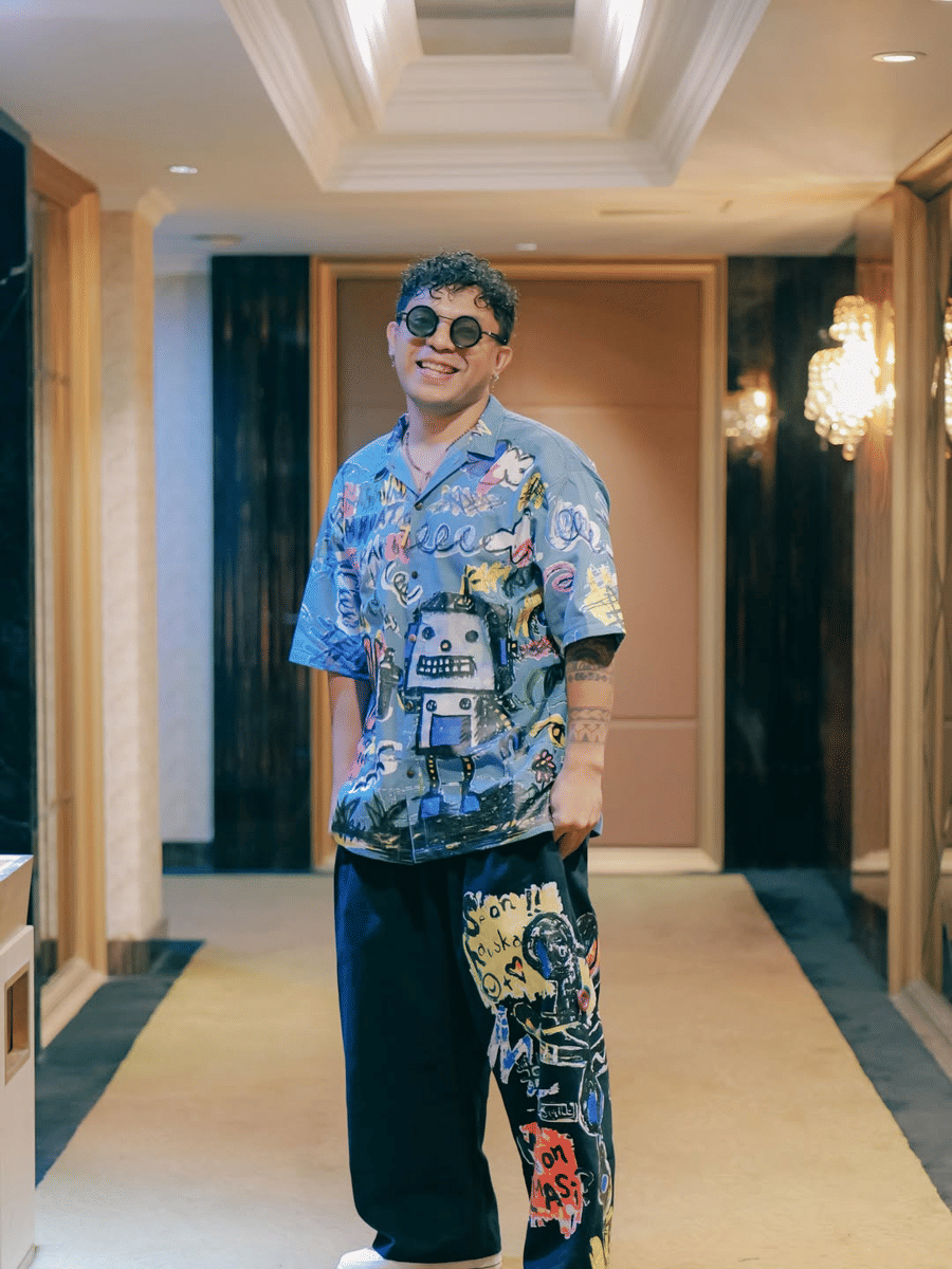 Silet Open Up mengenakan kemeja bermotif robot berwarna biru dan celana longgar bergambar seni jalanan berdiri di koridor hotel mewah.
