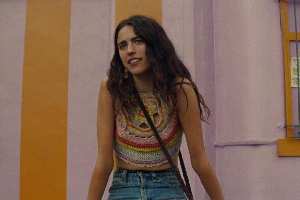 Margaret Qualley yang berambut panjang dengan atasan rajut warna-warni dan celana jeans berdiri di depan dinding bergaris oranye dan ungu.