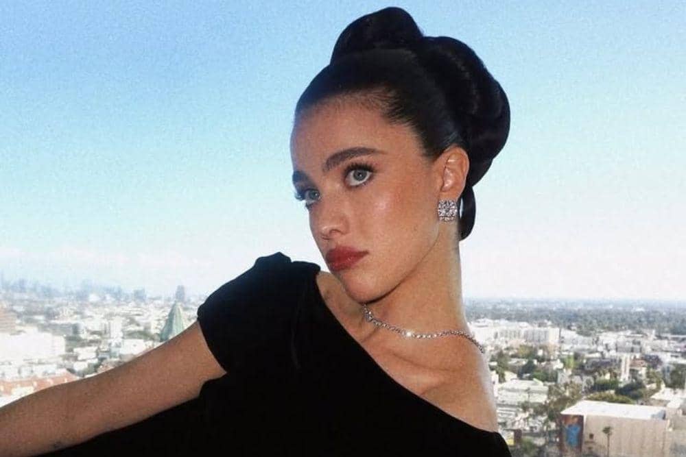 Margaret Qualley berpose elegan mengenakan gaun hitam dan perhiasan perak dengan latar belakang pemandangan kota di siang hari.