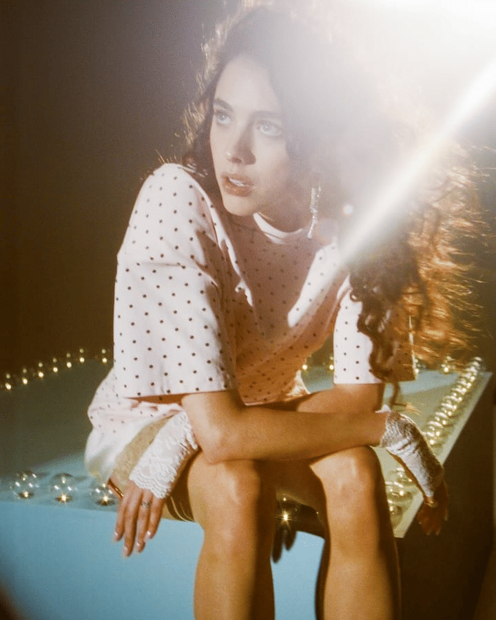 Margaret Qualley yang berambut panjang duduk dengan pencahayaan hangat, mengenakan blus putih bermotif polkadot dan sarung tangan renda.
