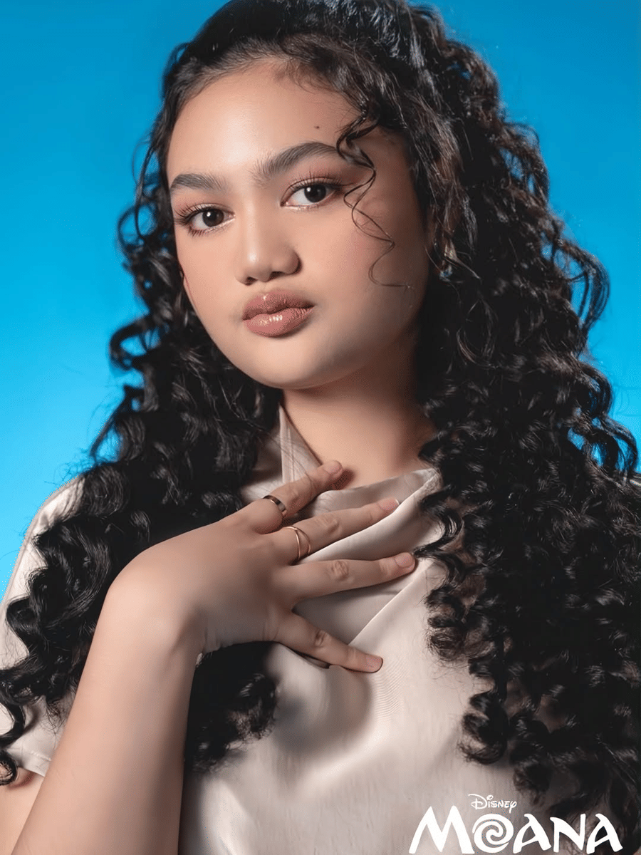 Amora Lemos yang berambut ikal panjang dengan pakaian satin berwarna terang berpose di depan latar biru, logo Disney Moana di sudut gambar.