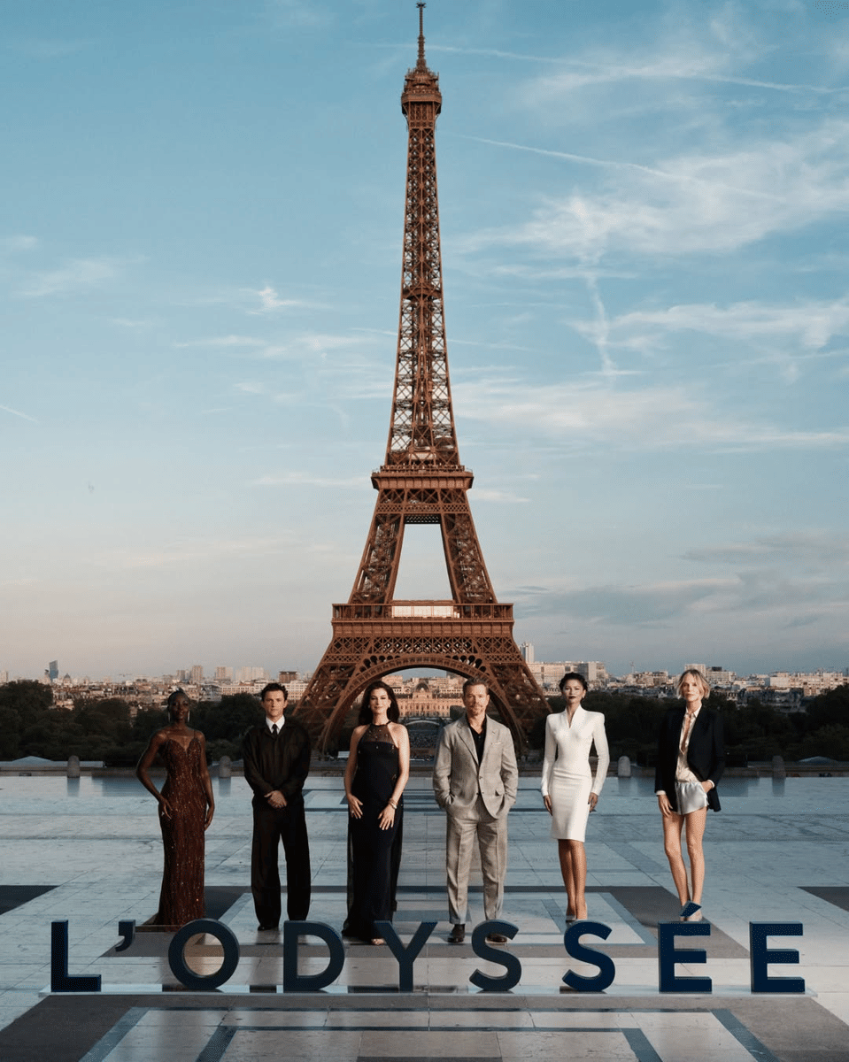 Para pemeran film The Odyssey berpose bersama di depan Menara Eiffel dengan latar langit biru dan tulisan L’Odyssée di bagian bawah.