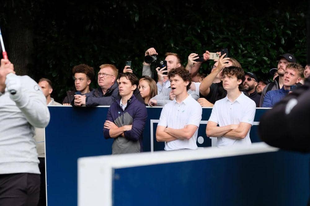 Tom Holland bersama kedua adiknya di tepi lapangan golf bersama penonton lain yang memotret dan menyaksikan turnamen di Wentworth Golf Club.