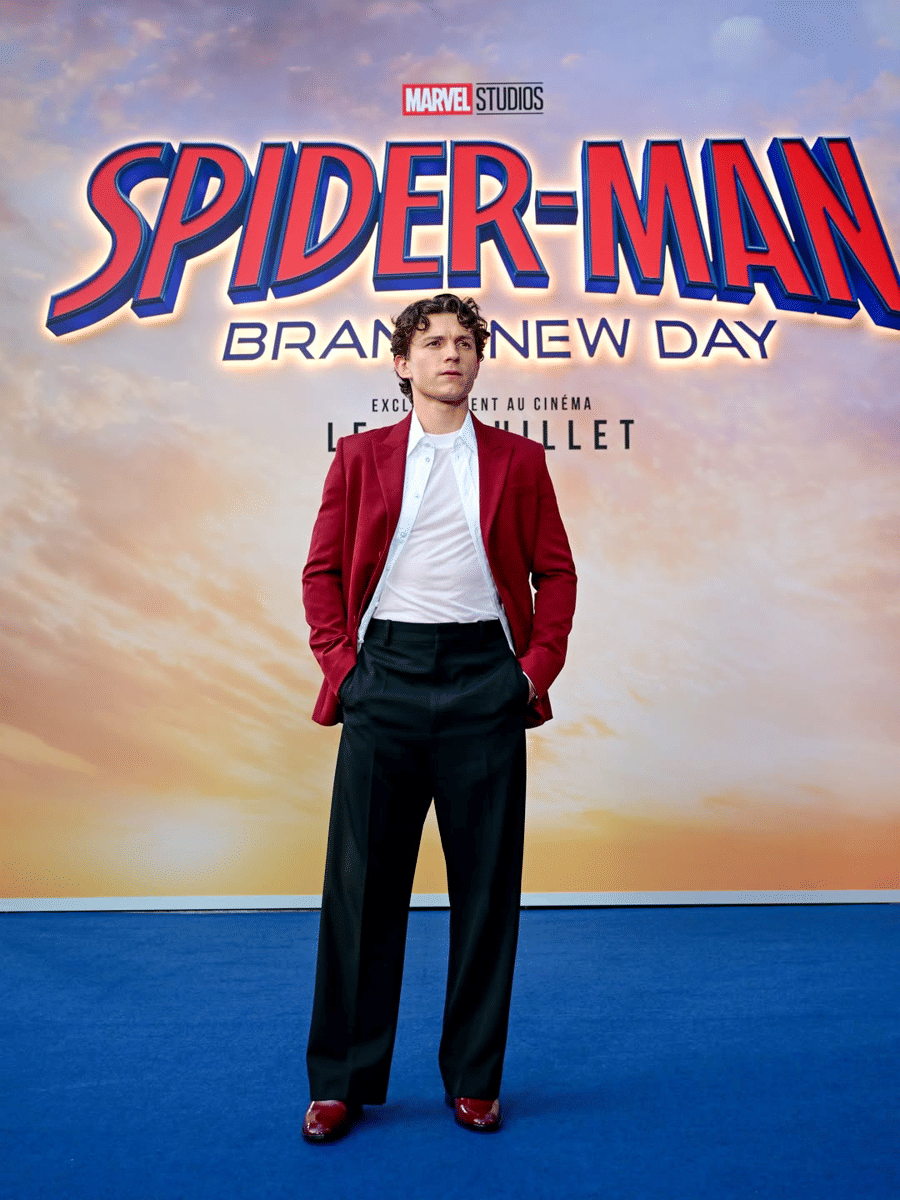 Tom Holland mengenakan jas merah dan celana hitam berdiri di depan latar bertuliskan Spider-Man: Brand New Day saat press tour di Paris.