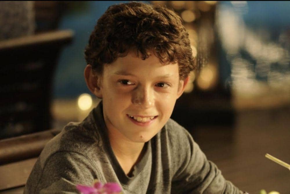 Tom Holland sebagai Lucas yang berambut ikal mengenakan kaus abu-abu tersenyum dengan latar cahaya hangat di sekitarnya.