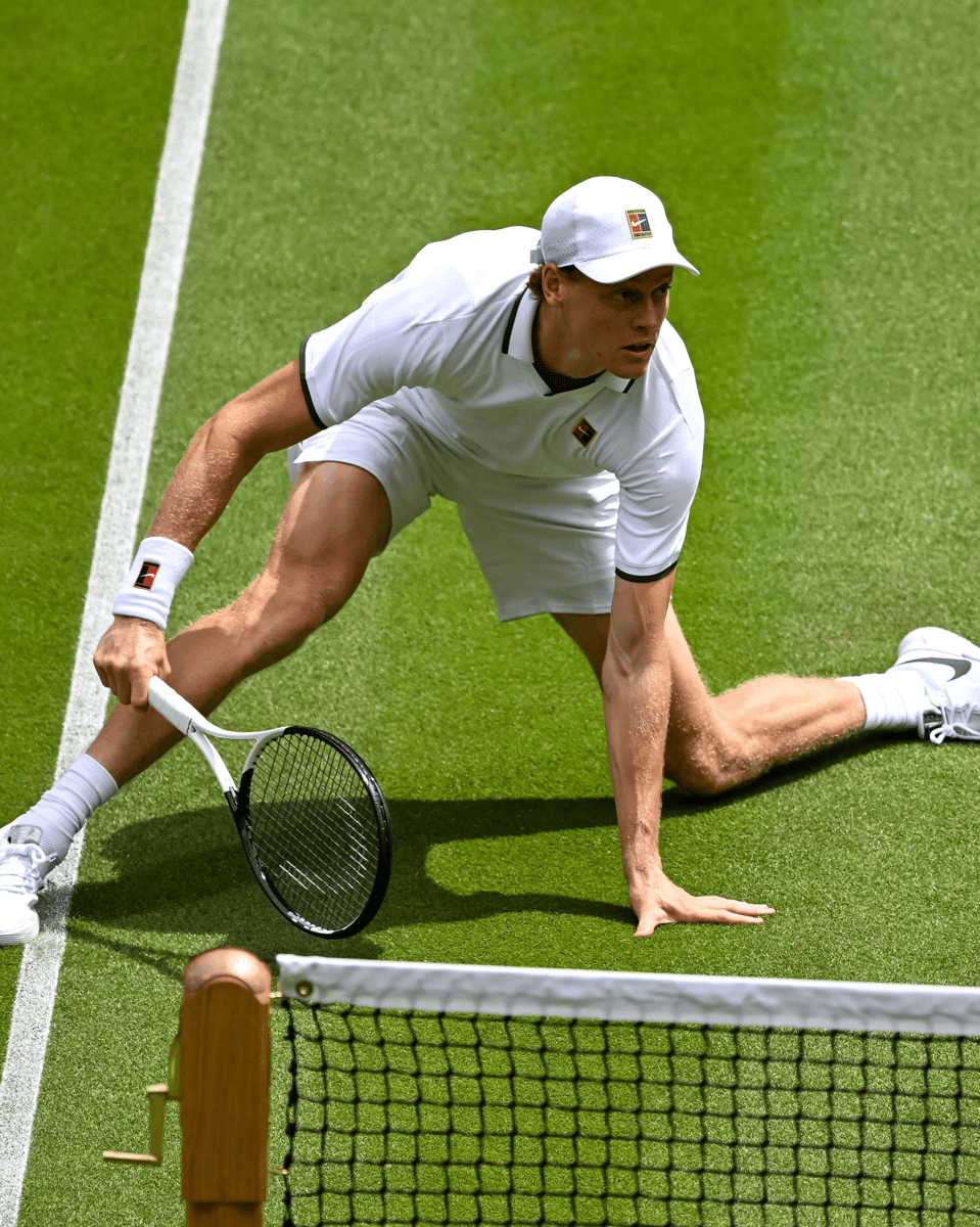Jannik Sinner mengenakan pakaian serba putih sedang beraksi di lapangan rumput Wimbledon dengan posisi rendah mendekati bola.
