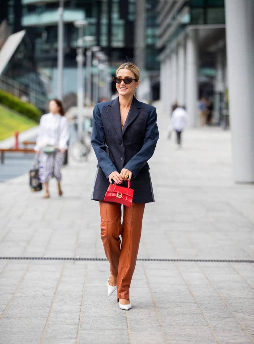 Seorang wanita bergaya mengenakan blazer biru tua, celana kulit cokelat, sepatu hak putih, dan tas kecil merah berjalan di area perkotaan modern.