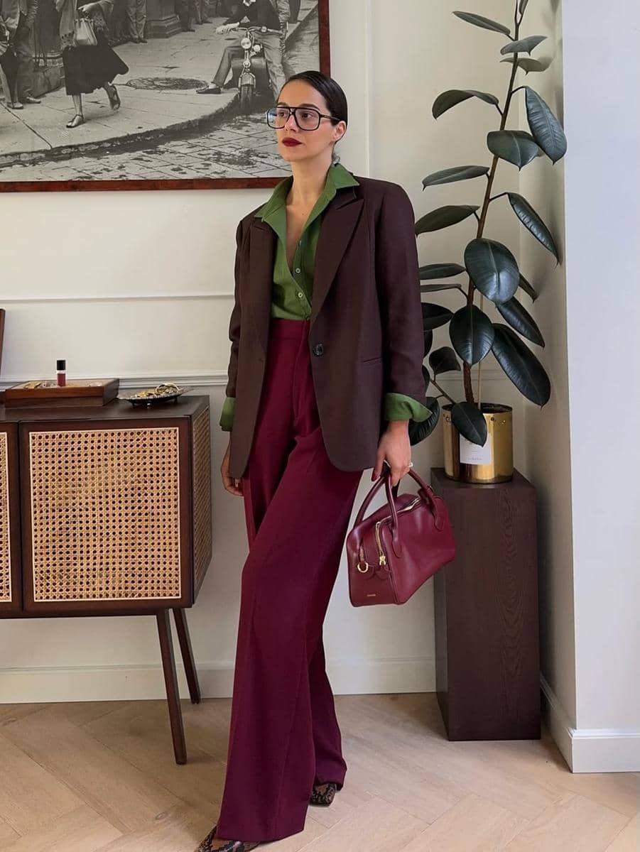 Seorang wanita bergaya elegan mengenakan blazer cokelat, kemeja hijau, celana panjang marun, dan membawa tas tangan merah di ruangan modern.
