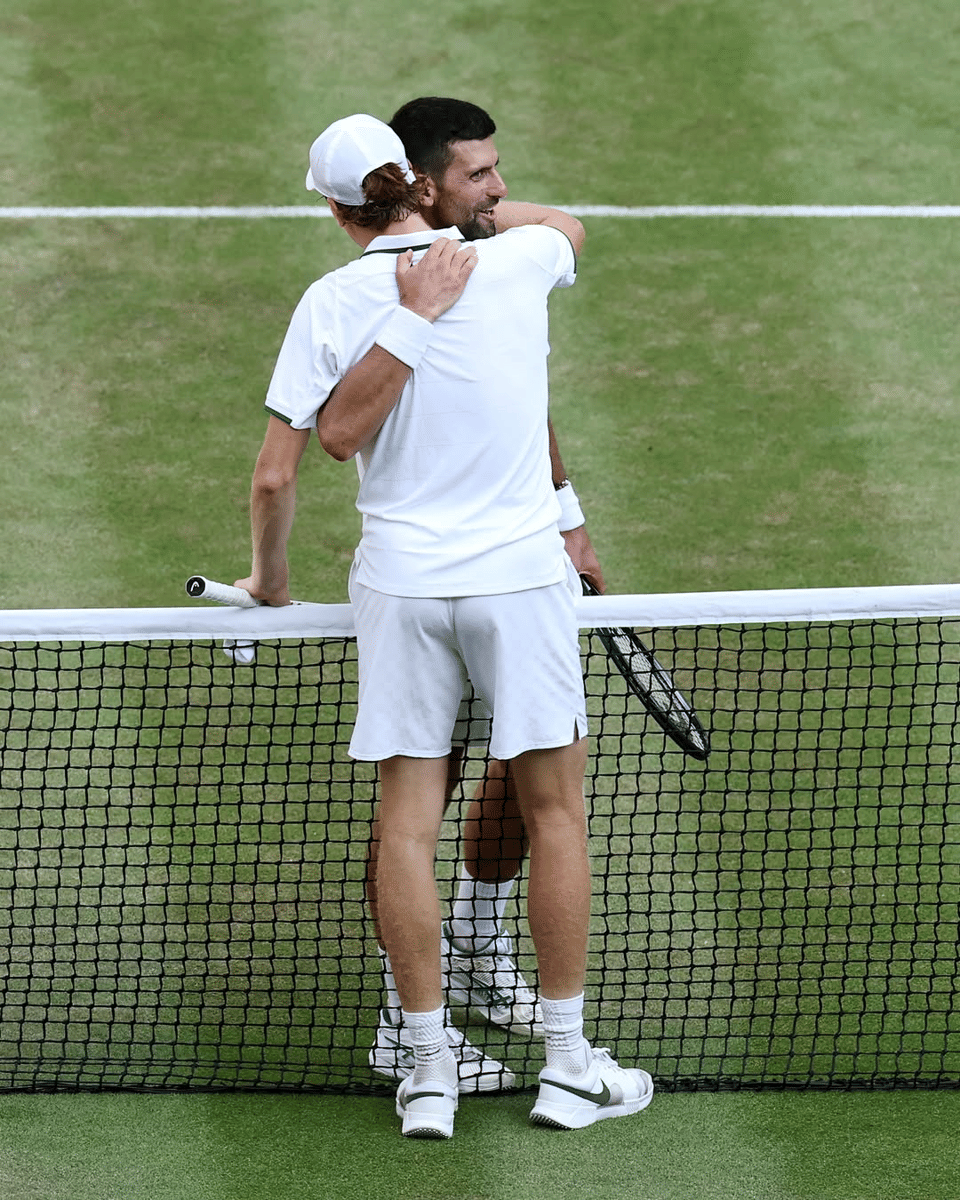 Dua petenis saling berpelukan di atas lapangan rumput Wimbledon setelah pertandingan, keduanya mengenakan pakaian putih dan memegang raket.