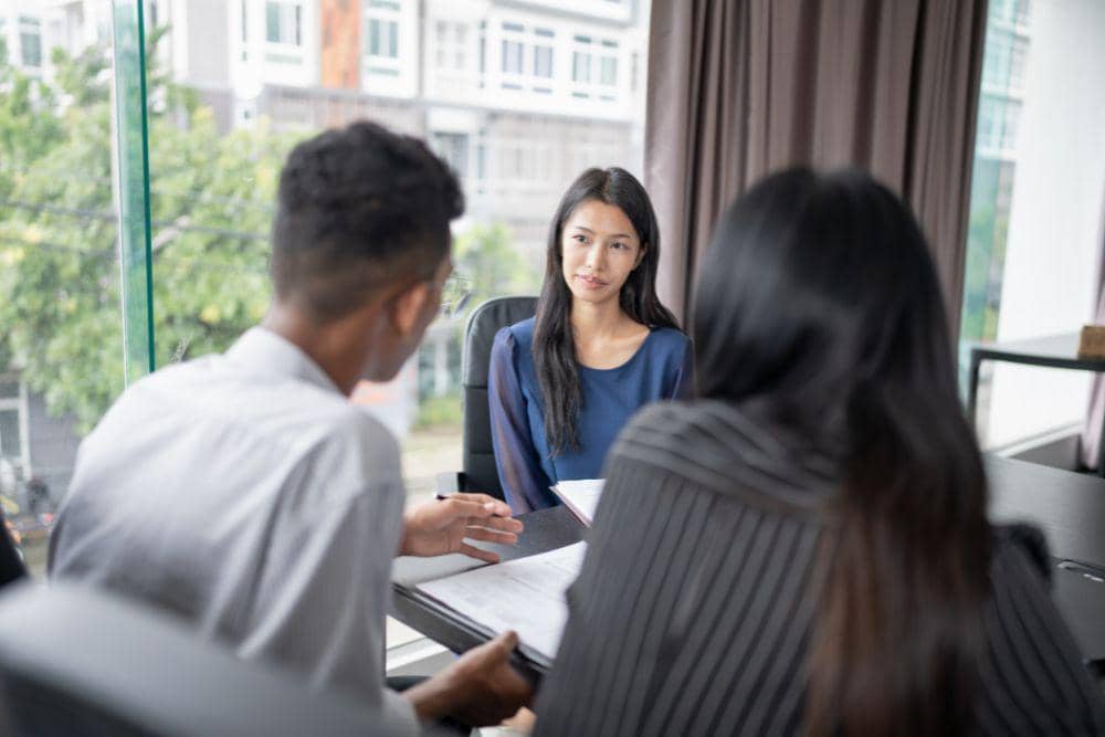Seorang wanita sedang menjalani wawancara dengan dua pewawancara di ruang kantor modern dengan jendela besar menghadap ke luar.