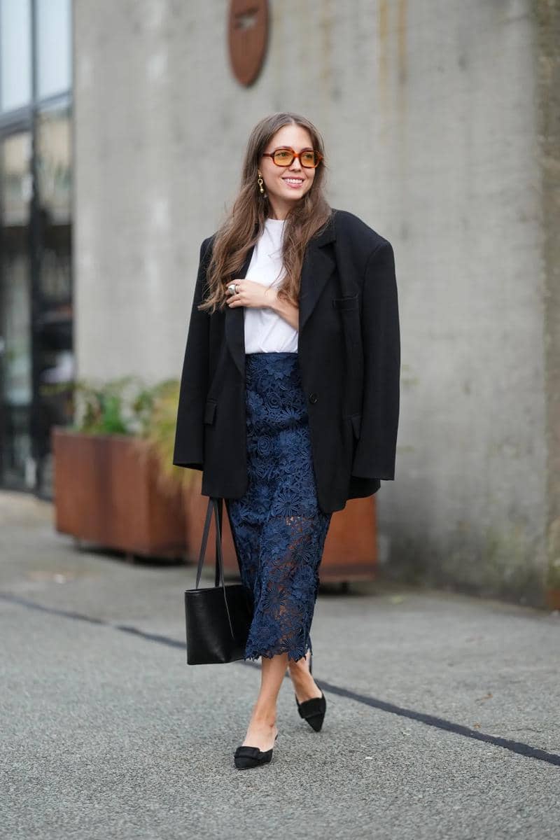 Seorang wanita berjalan di trotoar mengenakan blazer hitam longgar, kaus putih, rok renda biru tua, dan sepatu hitam dengan tas tangan hitam.