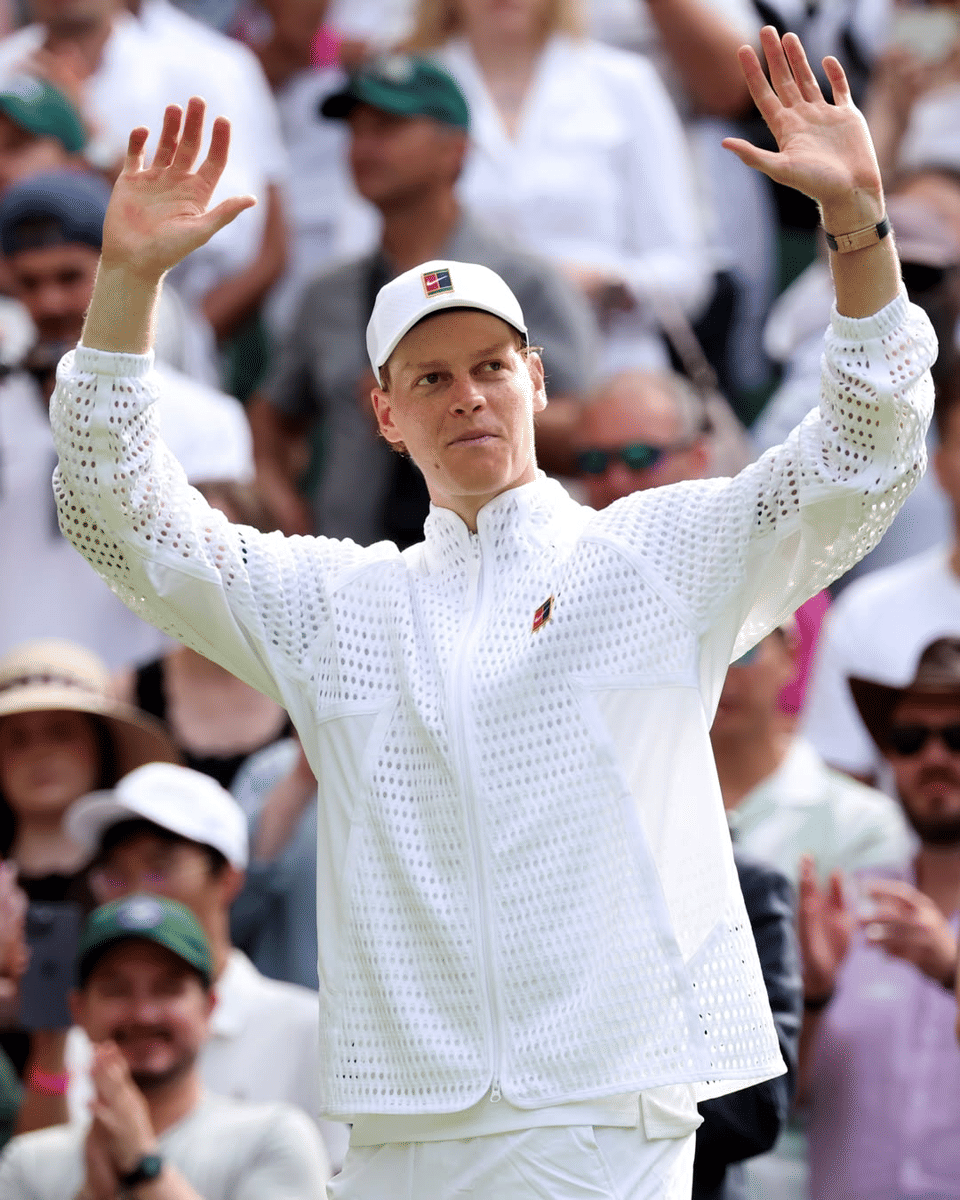 Jannik Sinner mengenakan pakaian putih lengkap dengan jaket dan topi, mengangkat kedua tangan di lapangan Wimbledon di depan penonton.