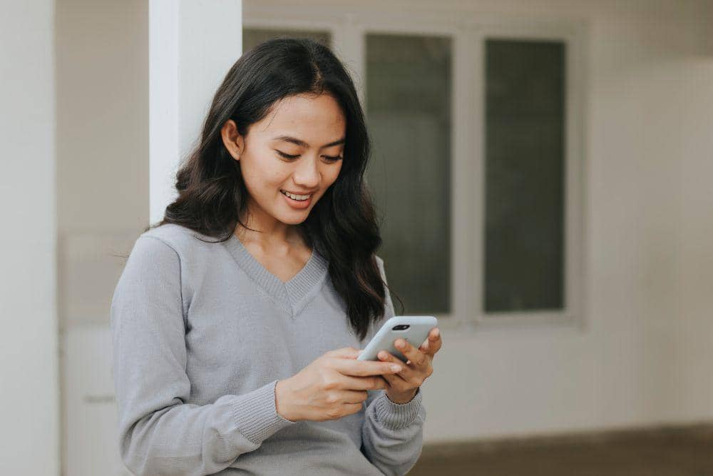 Seorang wanita muda mengenakan sweater abu-abu tersenyum sambil melihat ponsel di tangannya di dalam ruangan terang.