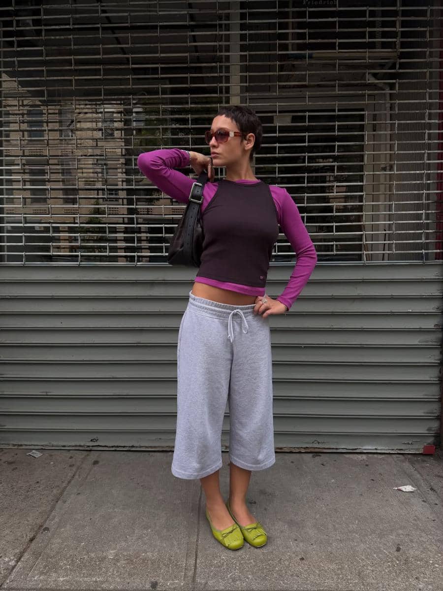 Seorang wanita bergaya kasual mengenakan atasan ungu dan hitam, celana pendek abu-abu, serta sepatu hijau di trotoar kota.