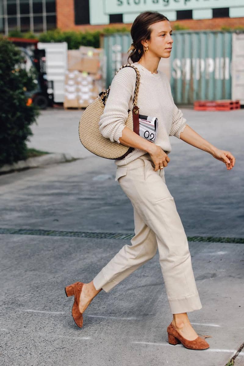 Seorang wanita berjalan mengenakan sweater krem, celana panjang beige, sepatu hak cokelat, dan tas besar anyaman di bahunya.