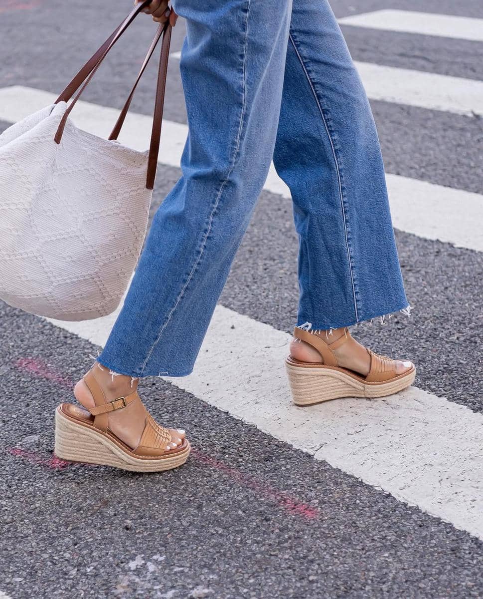 Seseorang berjalan di penyeberangan jalan mengenakan sandal wedge cokelat dan celana jeans biru dengan ujung terurai sambil membawa tas putih besar.