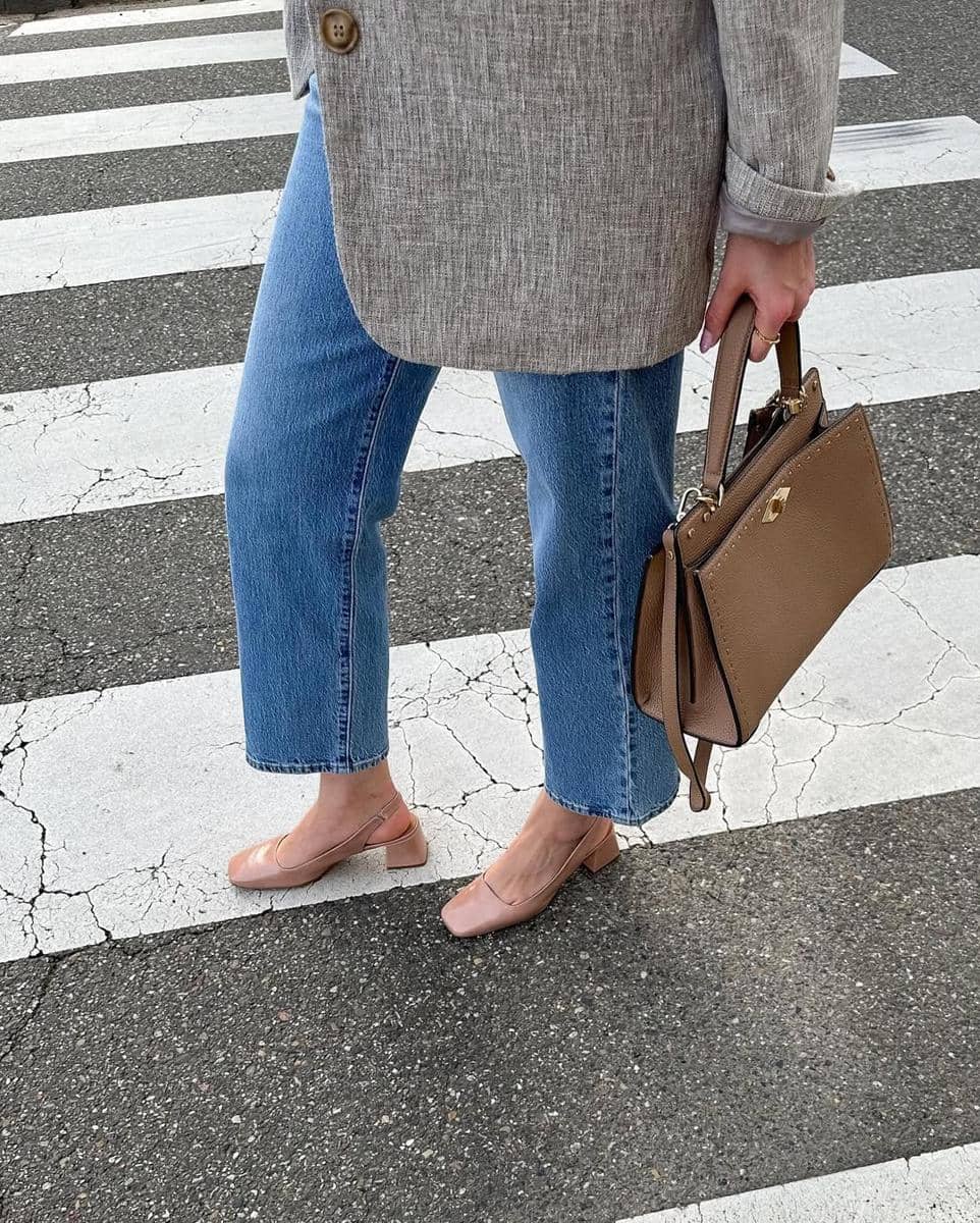 Seseorang mengenakan blazer abu-abu, celana jeans biru, dan sepatu hak rendah warna nude sambil membawa tas cokelat di penyeberangan jalan.