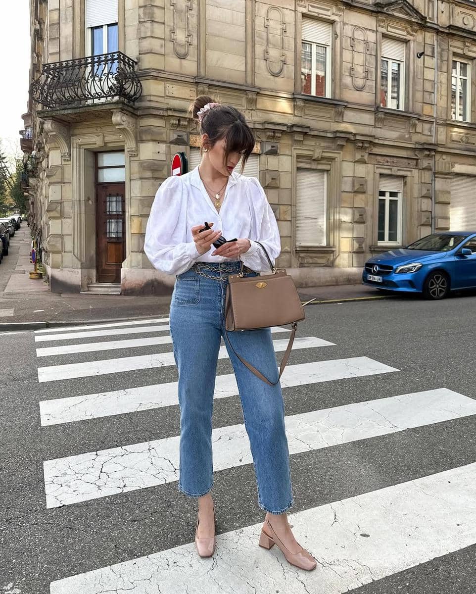 Seorang wanita berdiri di penyeberangan jalan mengenakan blus putih, celana jeans biru, dan sepatu hak rendah krem sambil memegang ponsel.