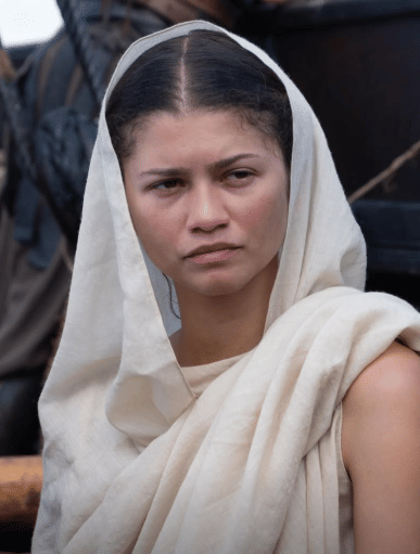 Seorang perempuan mengenakan kain berwarna krem menutupi kepala dan bahunya, berdiri di luar ruangan dengan latar perahu kayu.