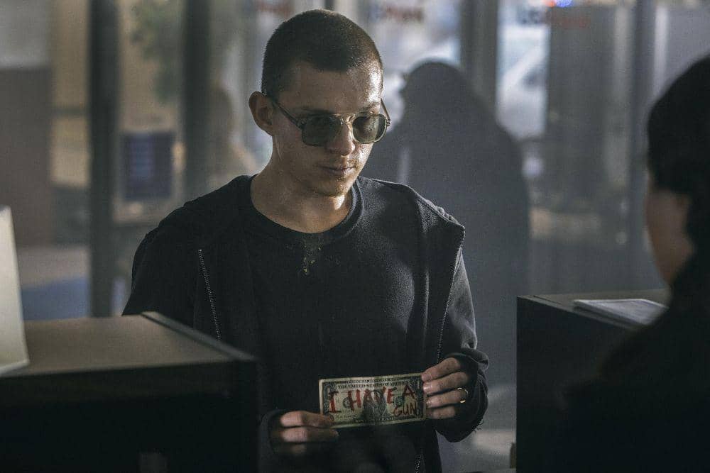 Tom Holland berambut pendek mengenakan kacamata hitam dan jaket gelap memegang secarik kertas di dalam ruangan bercahaya redup.