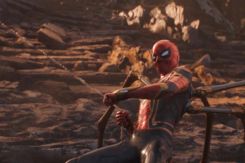 Spider-Man mengenakan kostum Iron Spider sedang menembakkan jaring di medan pertempuran berbatu dalam adegan film Avengers: Infinity War.