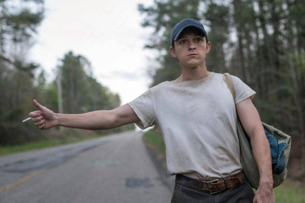 Tom Holland mengenakan kaus putih dan topi biru berdiri di tepi jalan sambil mengacungkan jempol untuk menumpang kendaraan.