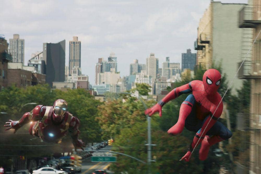 Spider-Man berayun di udara di antara gedung kota sementara Iron Man terbang di sampingnya dalam adegan film Spider-Man: Homecoming.
