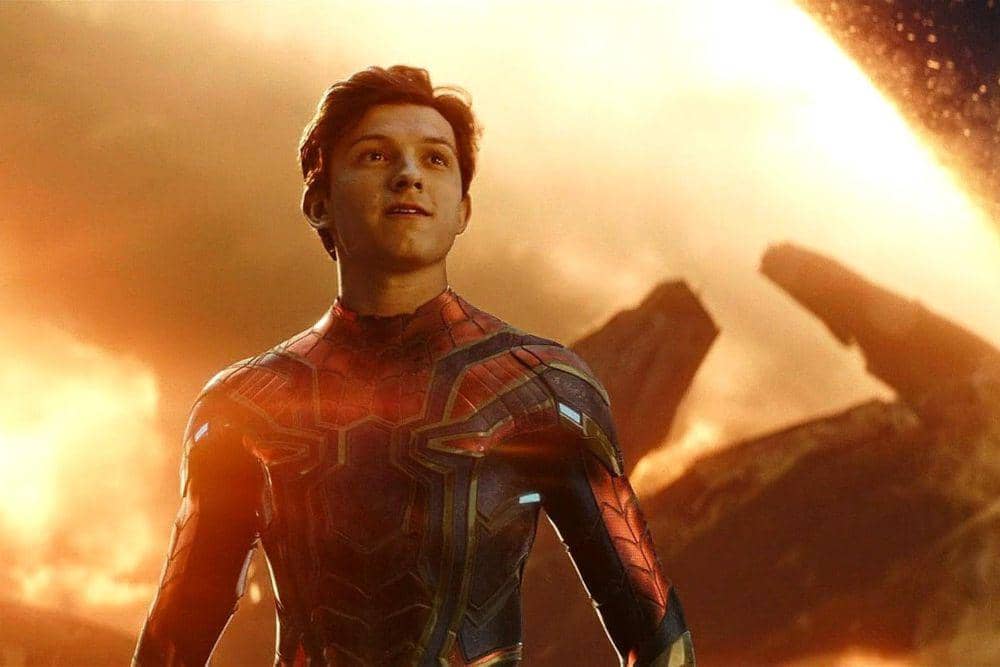 Tom Holland mengenakan kostum Iron Spider berdiri di latar belakang langit oranye dengan cahaya terang di sekitarnya.
