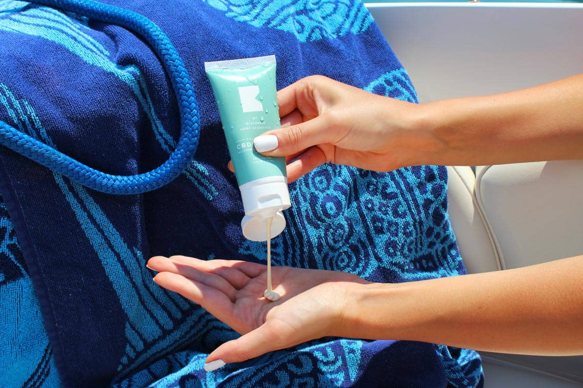 Seseorang sedang menuangkan produk sunscreen krim putih dari kemasan tube berwarna hijau toska ke telapak tangannya dengan latar belakang handuk pantai bermotif biru.