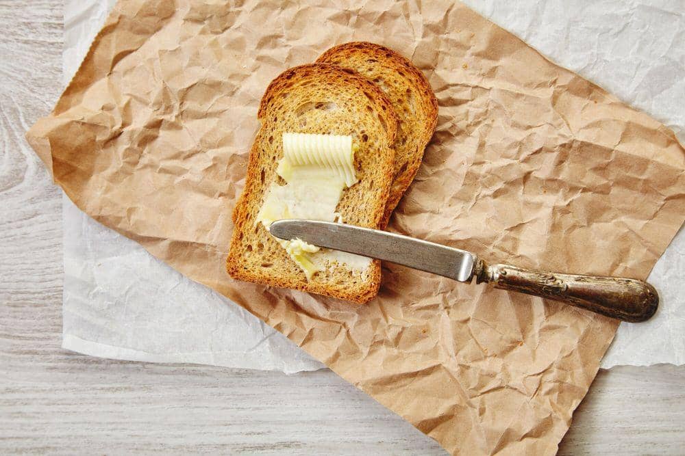Sepotong roti panggang berwarna cokelat keemasan diolesi butter lembut, disajikan di atas piring putih.